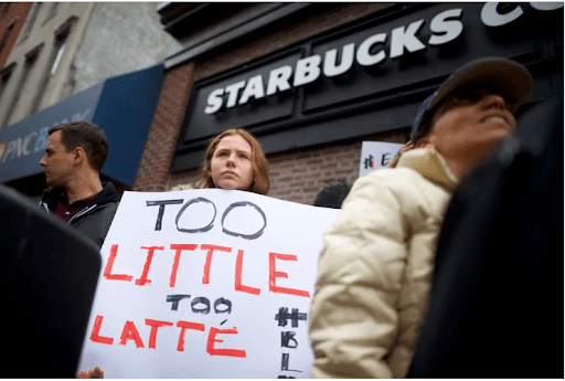 Woman protests outside starbucks with sign saying too little too latte